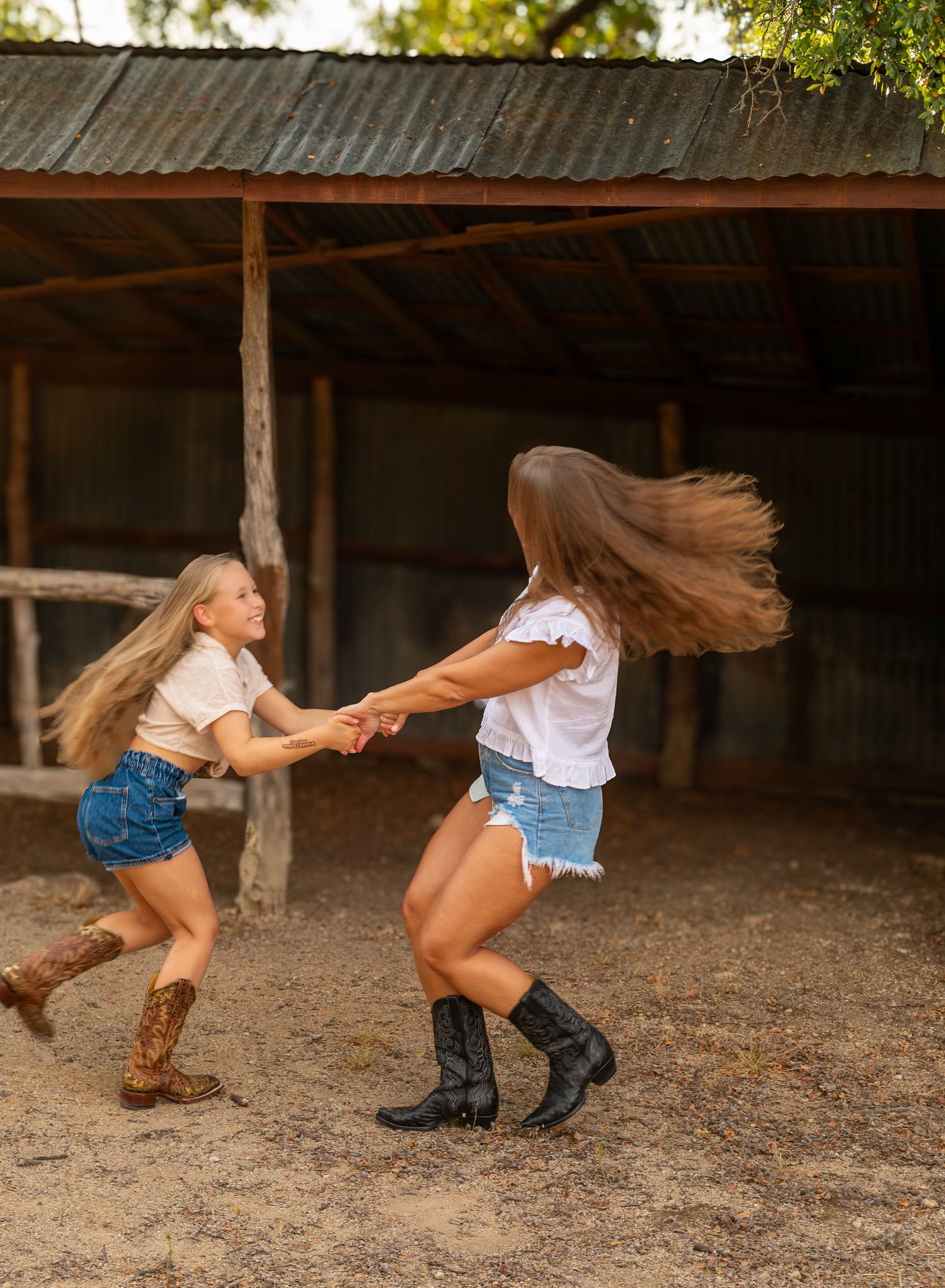Two friends in denim shorts and boots dance together on dirt ground under a rustic wooden shelter, their hair flowing in motion.
