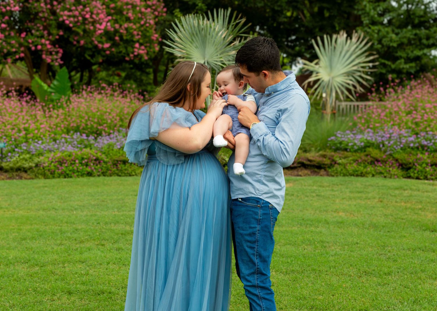 A couple in blue and denim embrace tenderly in a lush garden setting with blooming flowers and palm trees.