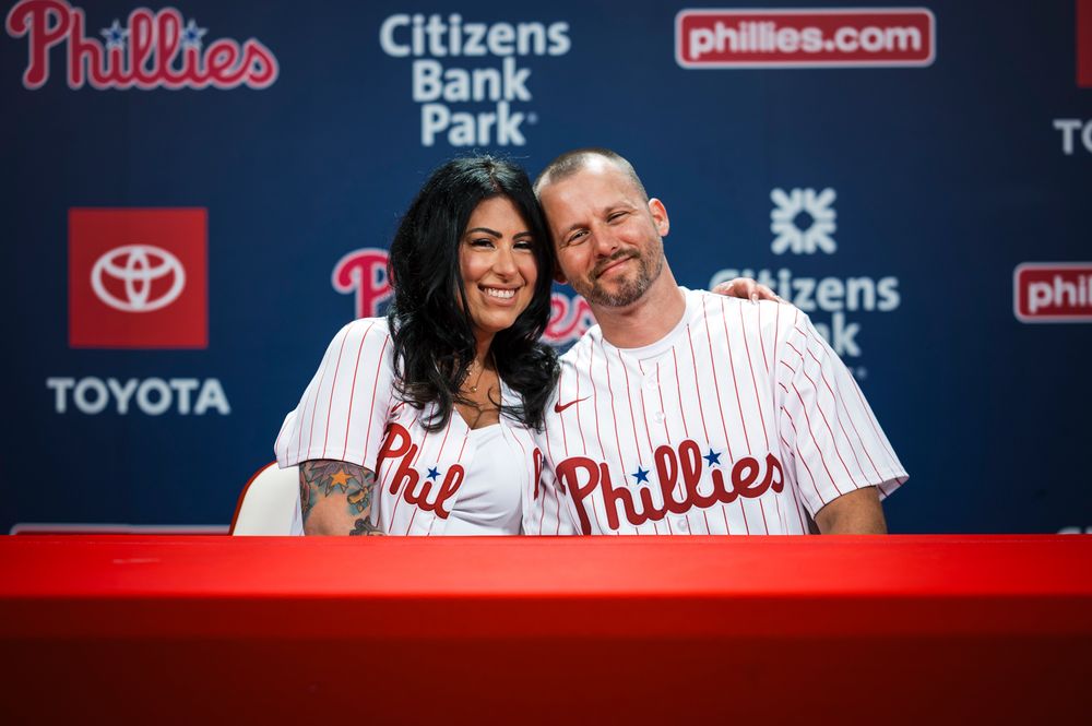 A couple in Phillies baseball jerseys smile and embrace while standing in front of a stadium display.