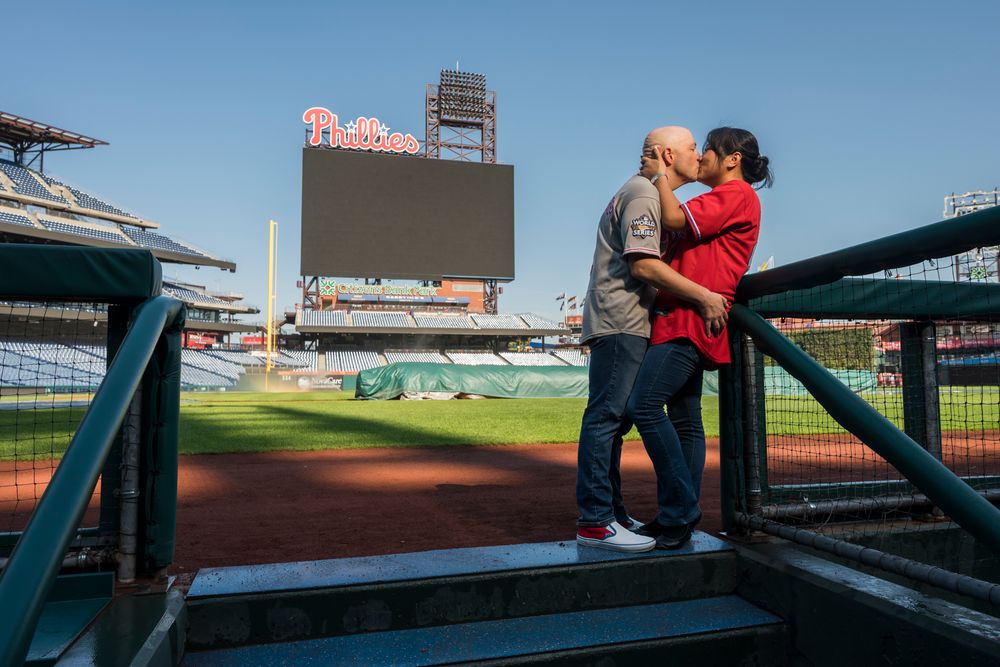 A couple embraces on the dugout steps of a baseball field Citizens Bank Park with the Phillies video scoreboard in the background