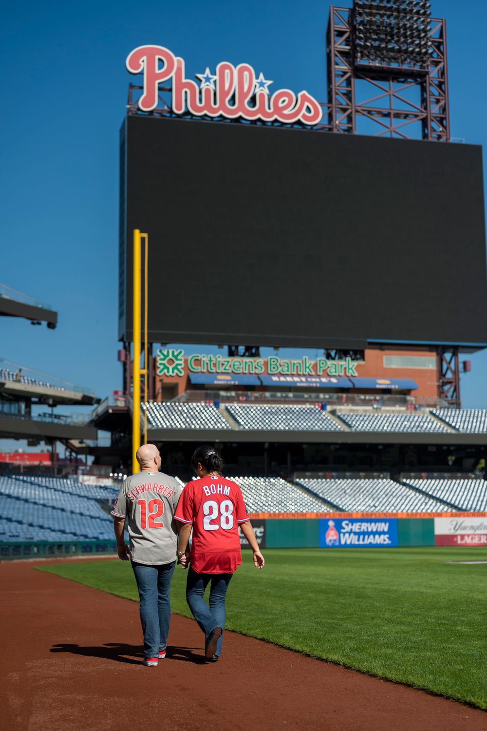 Two people walk along the warning track at Citizens Bank Park with the Phillies scoreboard visible in the background.