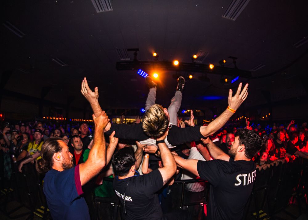 Concert crowd raises hands in silhouette against colorful stage lighting at a live music performance.