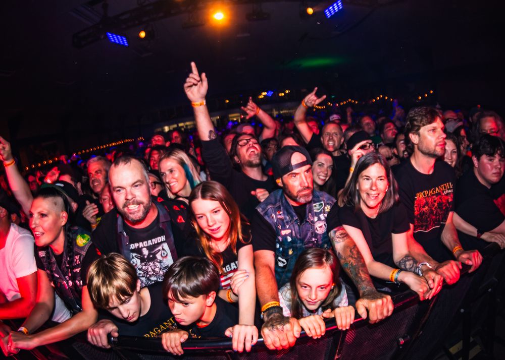 Energetic concert crowd cheering and raising hands at a dark venue with red and blue lighting.