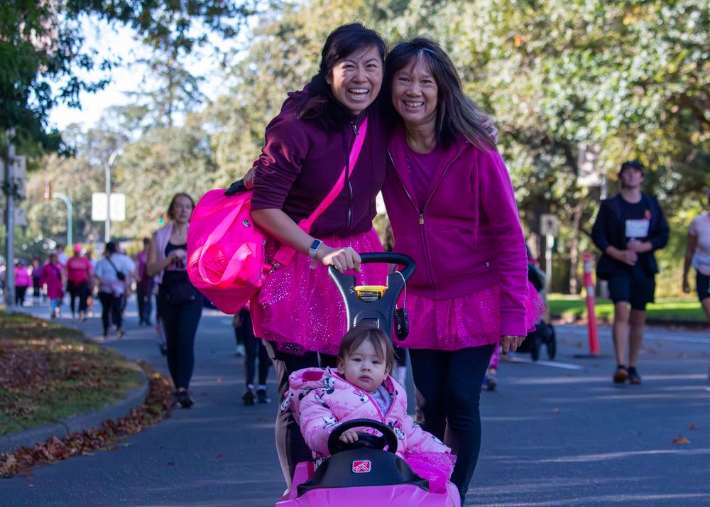 Participants in bright pink shirts run and walk together during an outdoor charity event on a tree-lined path.