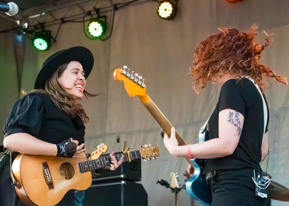 Musicians perform on stage with guitars and colorful stage lighting at an outdoor music festival.