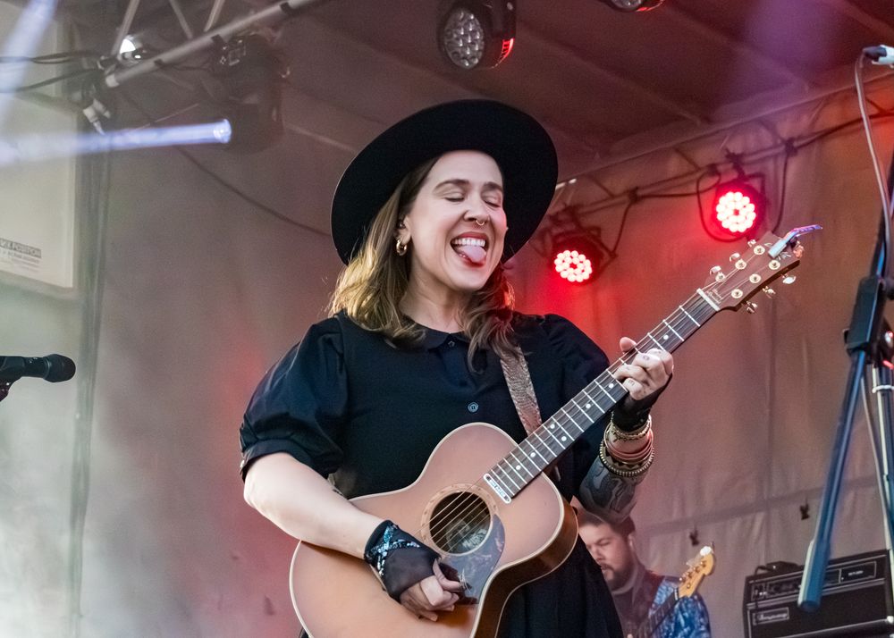 Musicians perform on stage with acoustic guitars and microphones under red stage lighting during a live concert performance.
