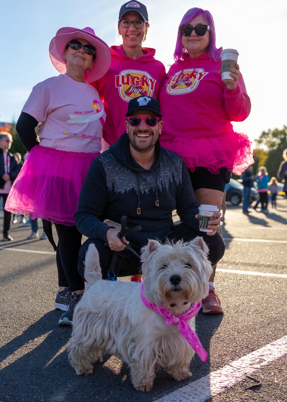 Group wearing pink outfits poses outdoors with a small white West Highland Terrier dog on a sunny day.