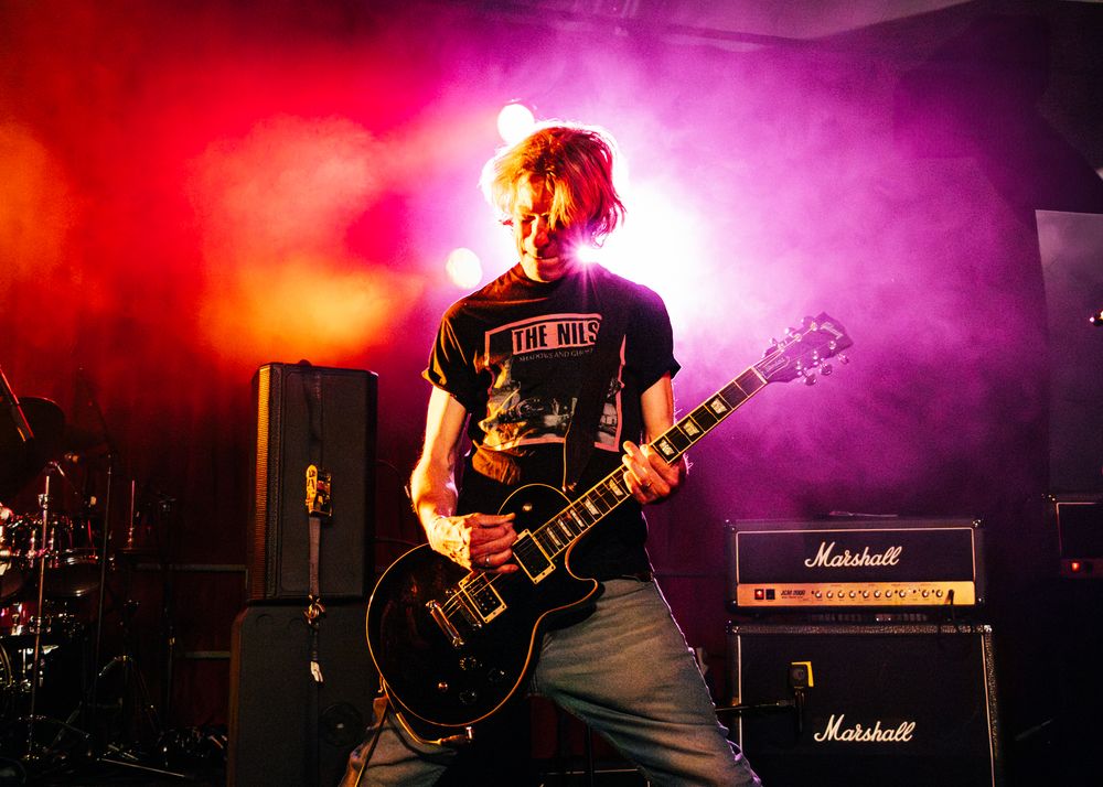 Rock band performs on stage with dramatic red and purple lighting behind Marshall amplifiers.