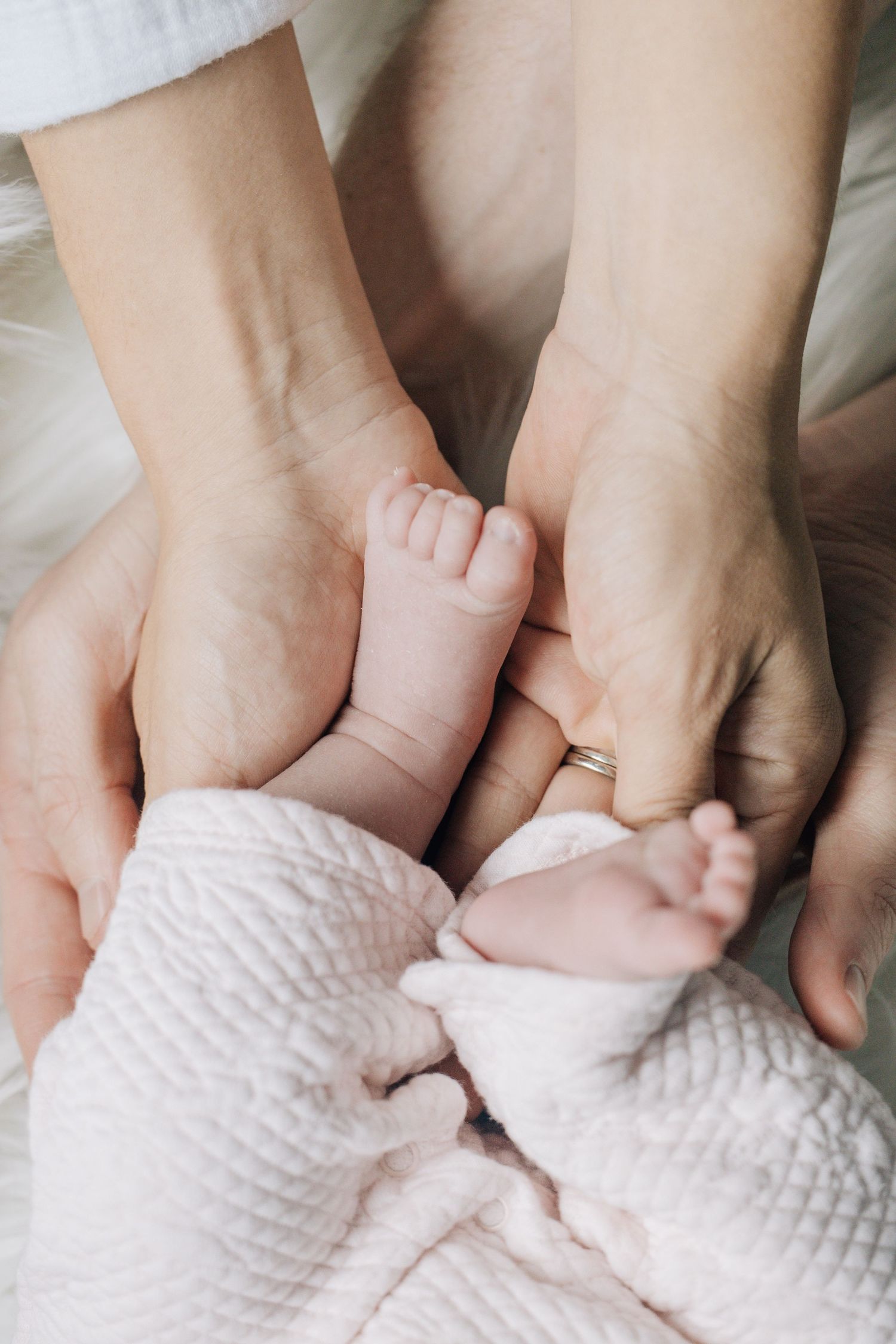 Adult hands gently cradling tiny newborn baby feet wrapped in a soft pink blanket.