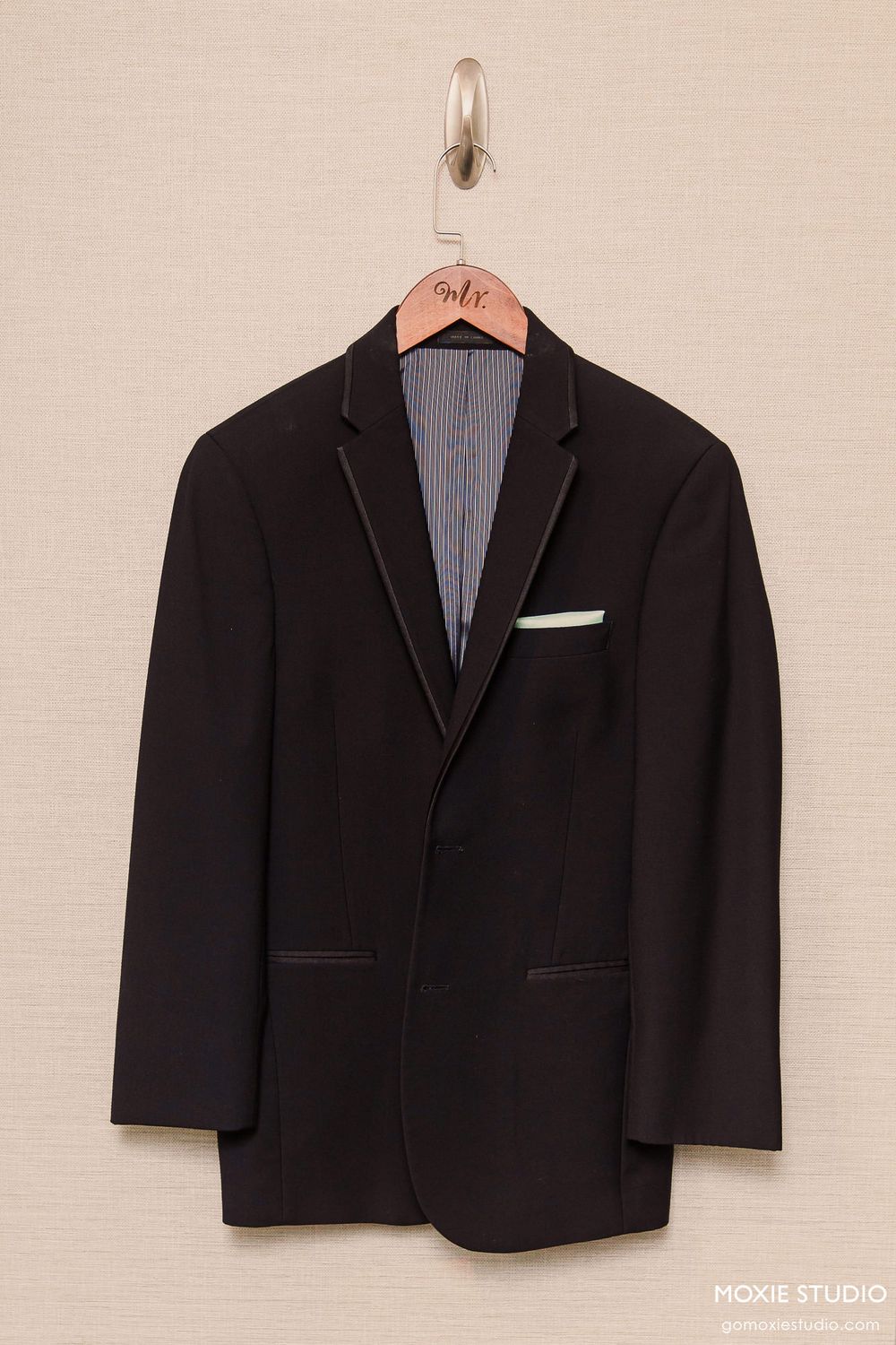 Black formal suit jacket displayed on a wooden hanger against a light colored wall.