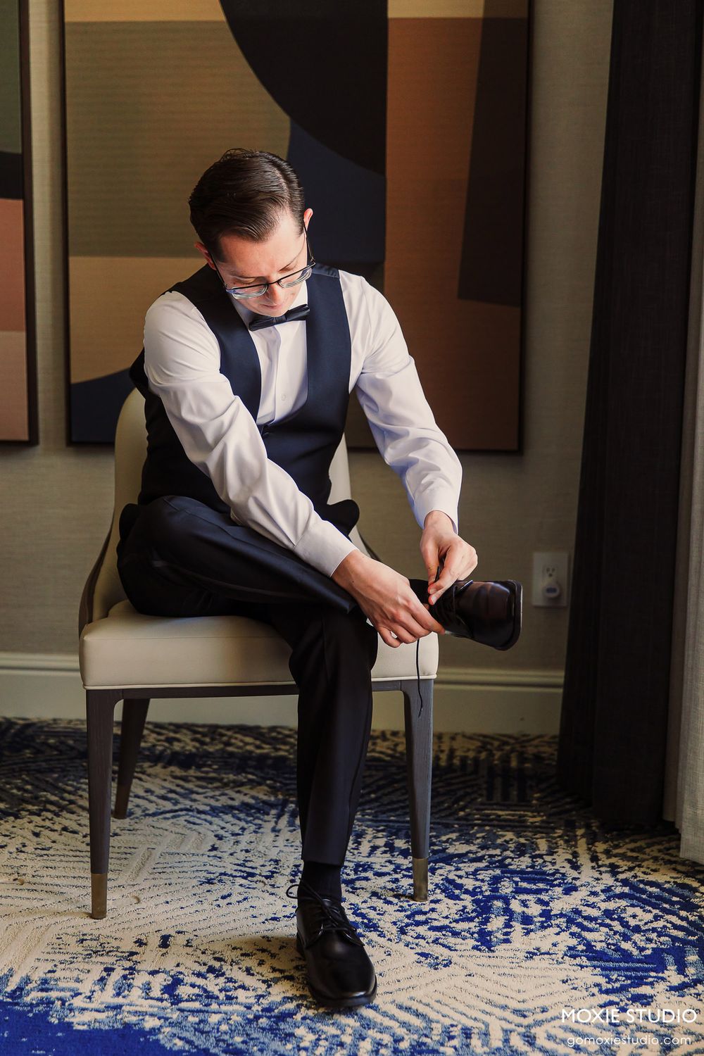 A groom in a navy suit and white shirt sits on a chair while getting ready on a patterned blue carpet.