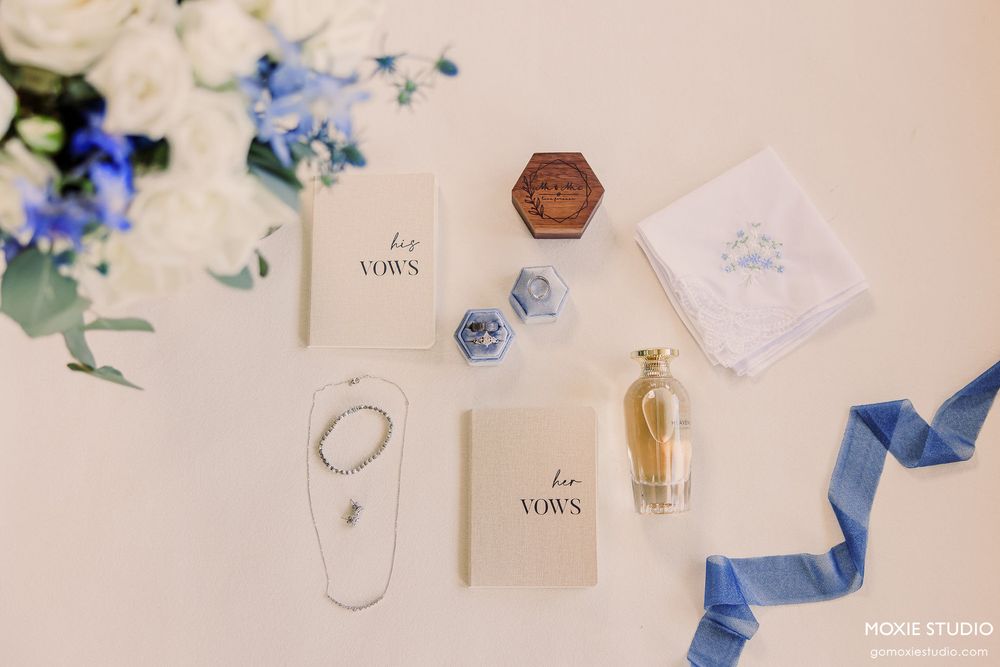 Wedding details flatlay with white and blue flowers, rings, perfume bottle, and blue ribbon on light surface.