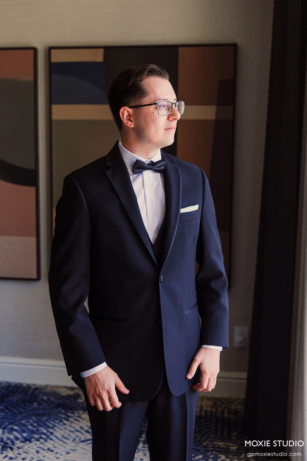 A groom in a classic navy tuxedo and bow tie stands in an elegant indoor setting with natural lighting.