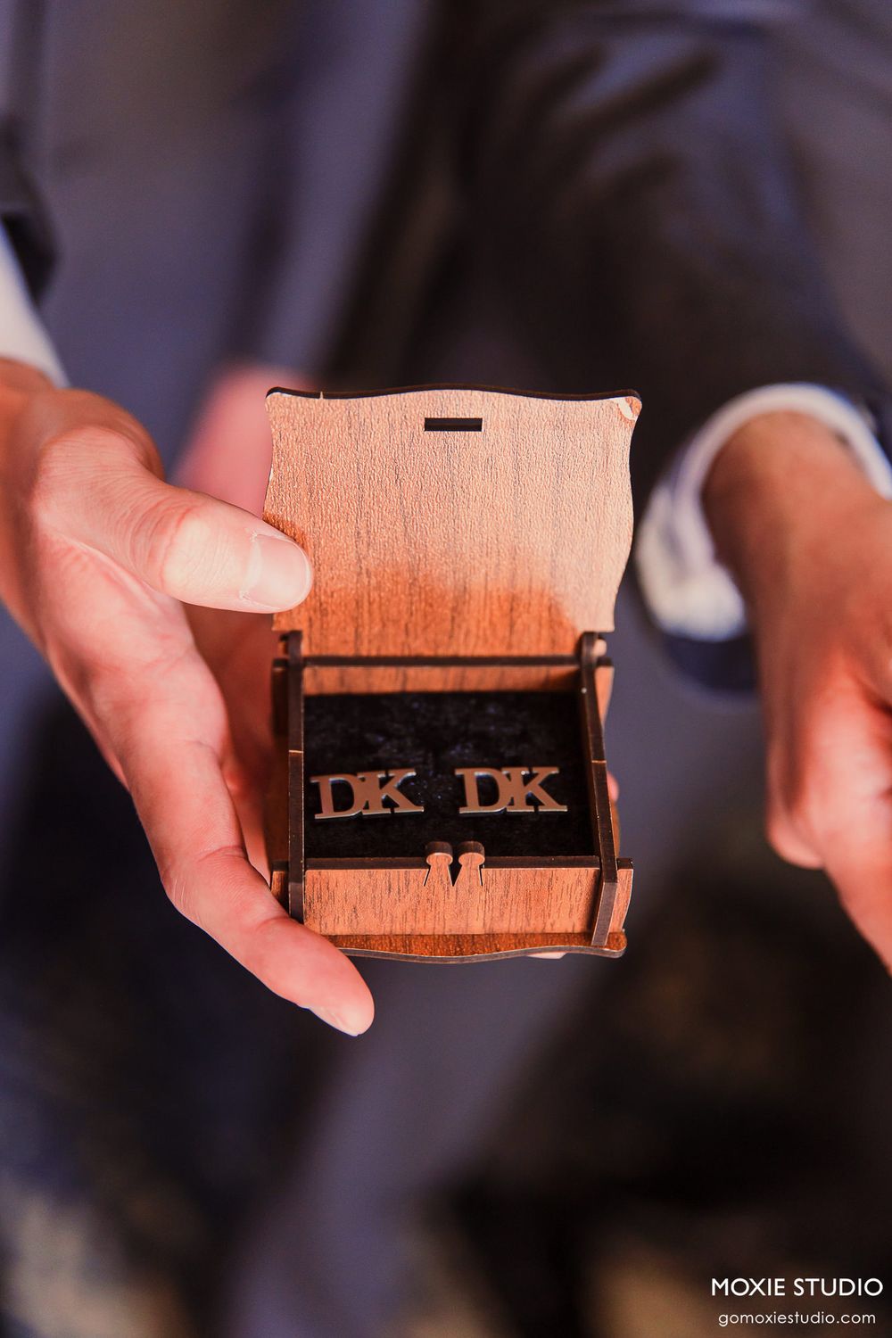 Hands holding and displaying a wooden box with DK letters engraved, shown in multiple angles against a dark background.