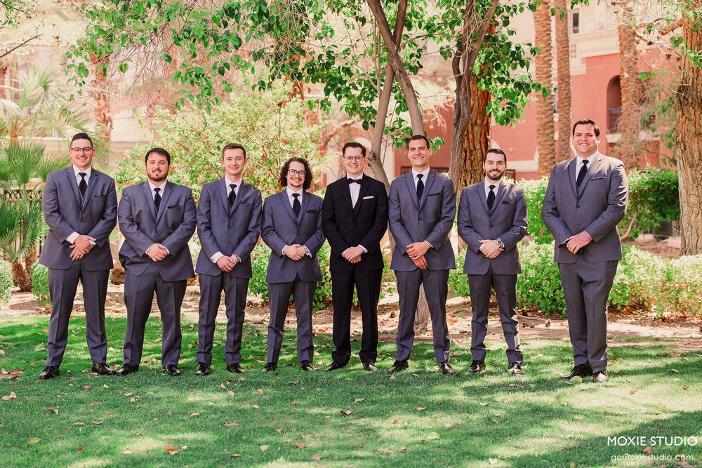 Wedding party groomsmen in matching navy suits pose together on green grass in front of a brick building.
