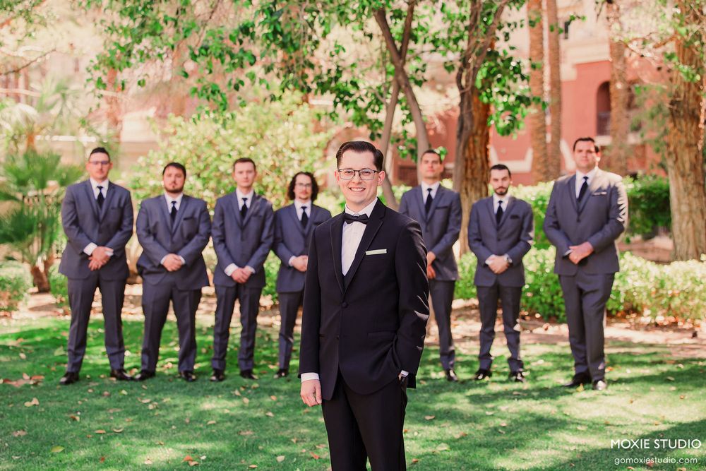 Wedding party in formal attire poses outdoors on a lawn with trees and brick building in background.