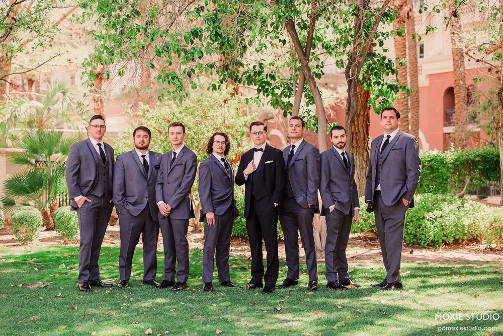 Wedding party in gray suits poses together on green lawn with pink stone building and trees in background.
