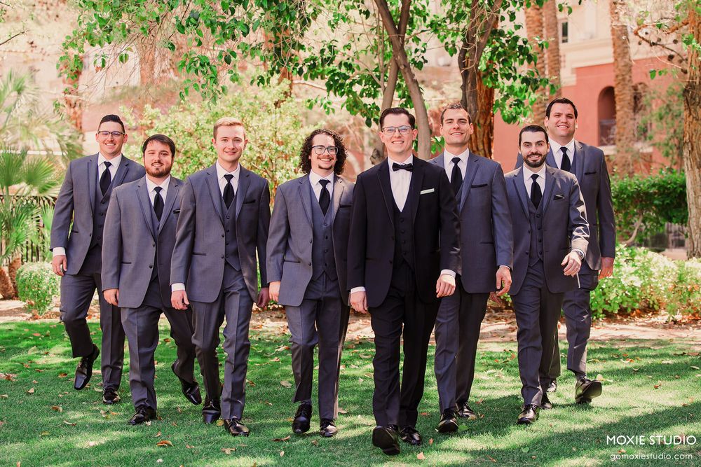 Groomsmen in matching charcoal suits walk in line on garden path with rustic pink wall backdrop.