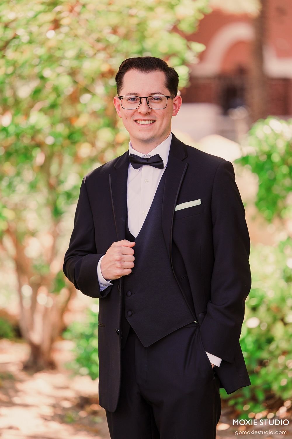 A groom in a classic black tuxedo poses outdoors against a garden backdrop with greenery.