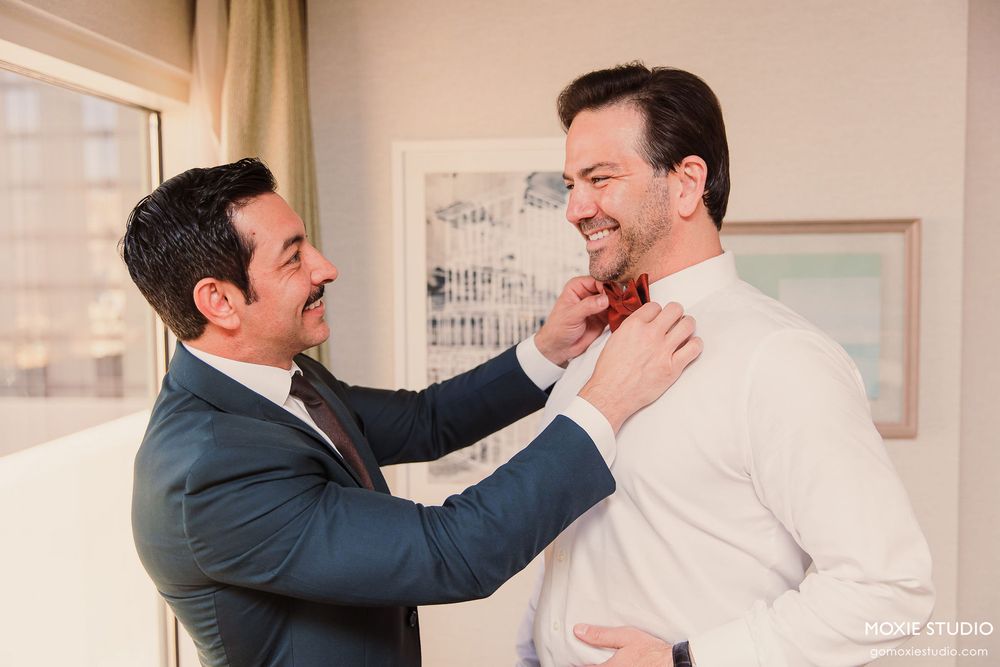 Two men in formal attire share a lighthearted moment while adjusting a bowtie before a wedding ceremony.