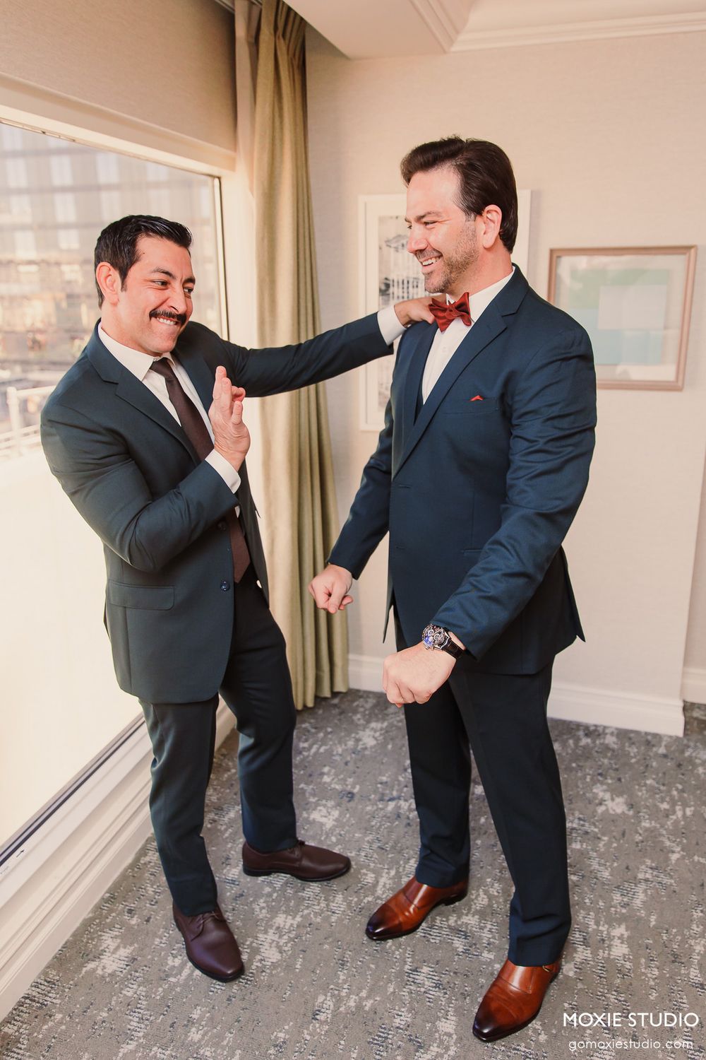 Two groomsmen in dark suits and bow ties share a playful moment before the wedding ceremony.