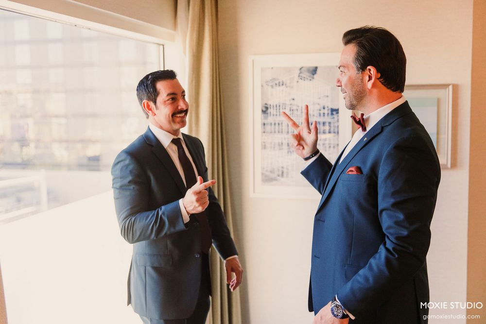 Two men in navy suits stand in conversation by a bright window.