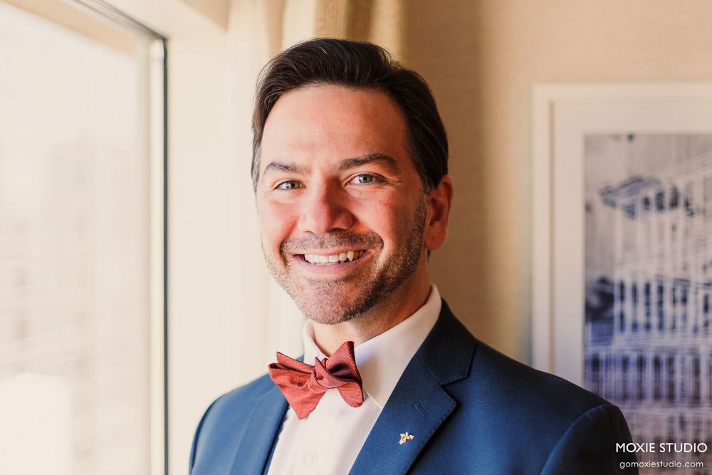 Groom in a navy suit and coral bowtie smiles warmly in natural window light.