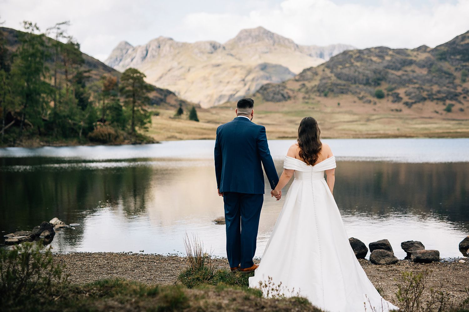 A couple in wedding attire stands at the edge of a serene mountain lake with majestic peaks in the background.