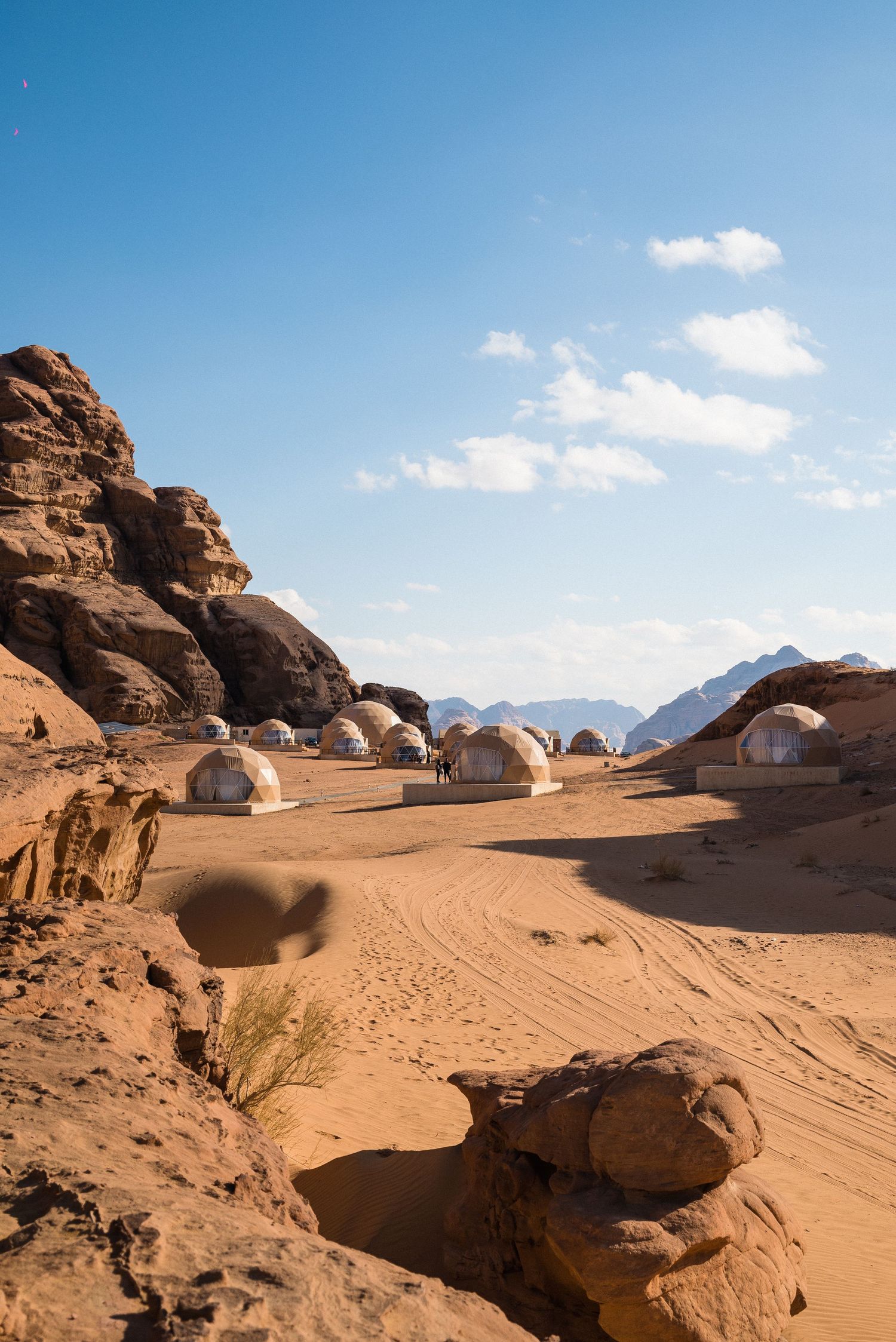 Desert camp with dome tents nestled between red rock formations under blue sky.