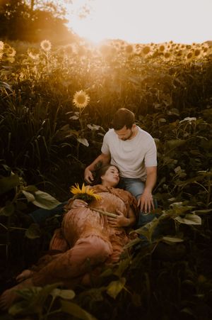 A romantic couple rests together in a sunlit sunflower field during golden hour as sun flares illuminate the scene.