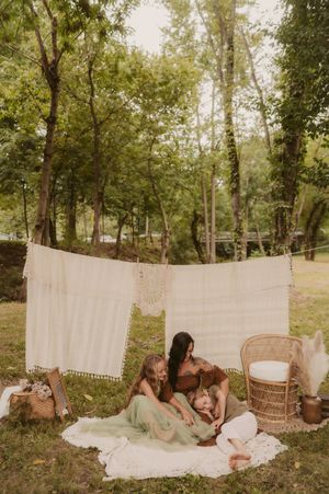 A dreamy outdoor scene with white linens hanging between trees while figures relax on blankets in a natural setting.