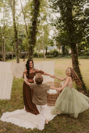 Three people in flowing dresses and casual wear dance together on a white blanket in a sunlit woodland setting.