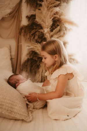 A young girl in a white dress sits on a bed with a newborn baby, surrounded by soft pampas grass decor.