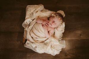 Overhead view of sleeping newborn wrapped in soft beige fabric on textured lace blanket against dark wooden floor.