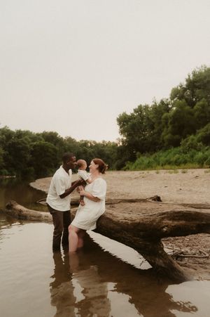 A couple in white clothing shares intimate moments by a fallen tree trunk near the water's edge at sunset.
