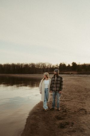A couple walks along a serene beach at sunset with a lake and treeline in the background.