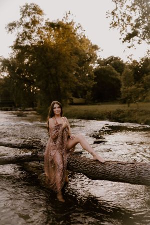A person in a floral dress poses on a fallen log by a river during golden hour at sunset.