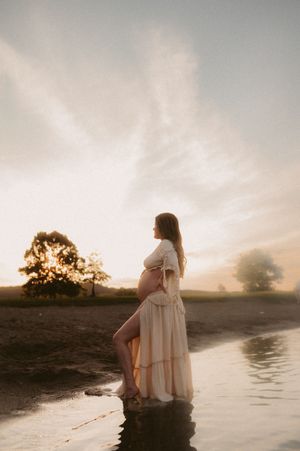 Scenic sunset maternity photoshoot by a tranquil lake showing silhouette of pregnant woman in flowing dress.