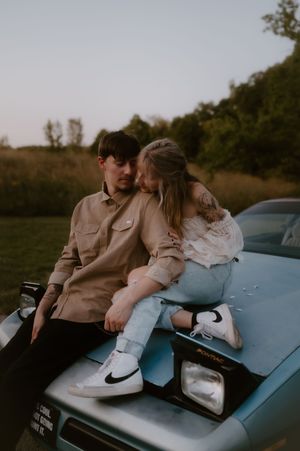 A couple in casual attire sits intimately on the hood of a light blue classic car at sunset.