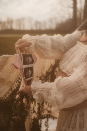 Someone in a white lace dress holds an ultrasound photo outdoors during sunset in a dreamy, vintage-style photograph.