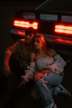 A couple sits in a dimly lit car interior illuminated by red ambient lighting and neon reflections.