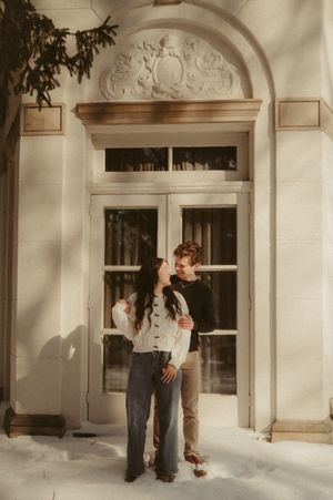 A couple stands together in front of an elegant arched doorway with ornate architectural details in warm sunlight.