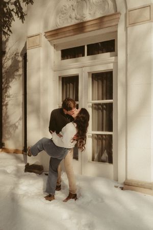 A person in grey clothing peeks through a window of a white building surrounded by snow.