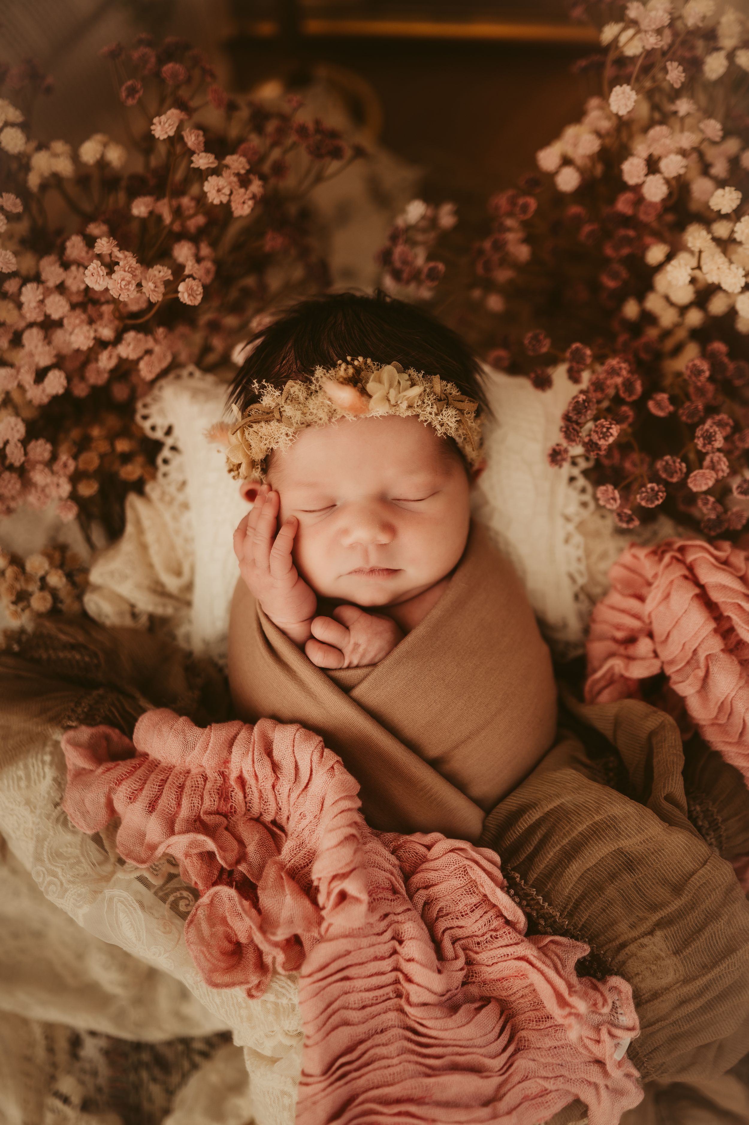 Sleeping newborn baby wrapped in tan swaddle with floral headband surrounded by pink flowers and ruffled pink blanket.