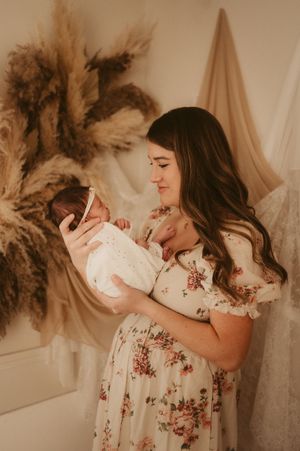 A young mother in a floral dress tenderly holds her newborn baby in a soft, bohemian-styled photo session.