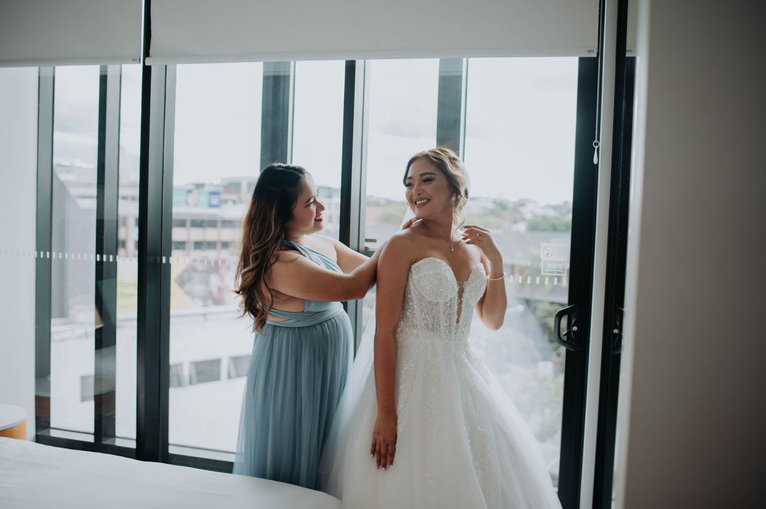 A bridesmaid helps adjust the wedding gown in a bright room with city views.