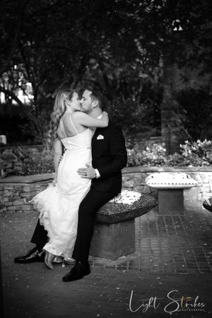 Black and white wedding photo of couple sharing romantic kiss while sitting on stone wall in garden setting.
