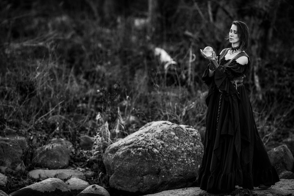 A ethereal figure in a gothic black dress performs mystical gestures near a large stone in a moody forest setting.