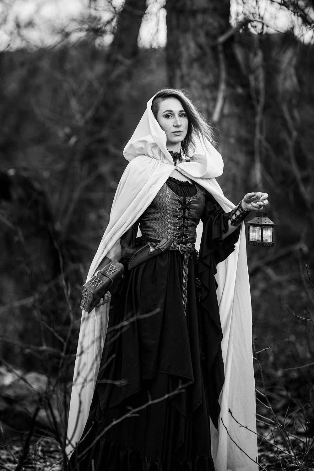 A dramatic black and white image shows a figure in medieval dress with corset and white cloak holding a lantern in dark woods.