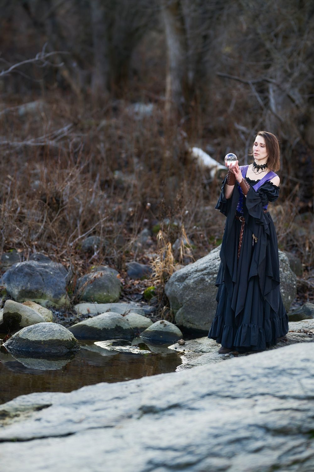 A figure in a flowing black dress stands on rocks by a stream in a moody forest setting, gesturing outward.